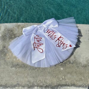 May include: White tulle skirt with a white satin bow and red glitter lettering that says "Future Mrs. Rojas".