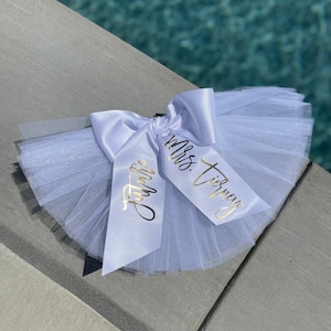 May include: White tulle bow with two white satin ribbons. The ribbons have gold lettering that says "Mrs. Tierney" and "Just Married".