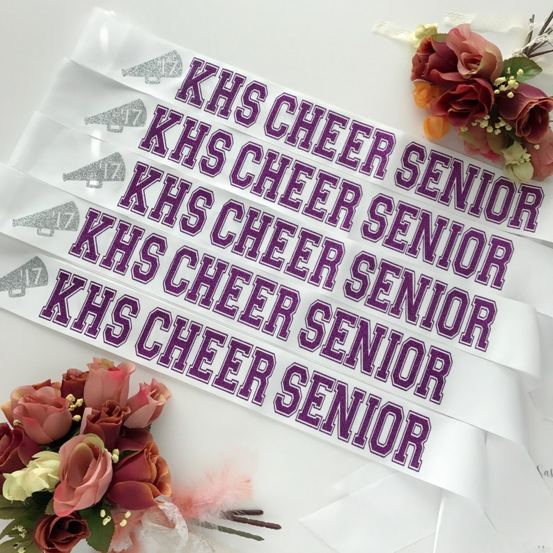 May include: Five white satin sashes with purple text that reads "KHS CHEER SENIOR". Each sash has a silver glitter megaphone with the number 17 on it.