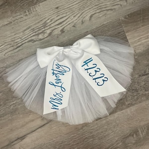 May include: White tulle tutu with a white satin bow and the text "Mrs. Lovely" and "12.3.23" in blue glitter.