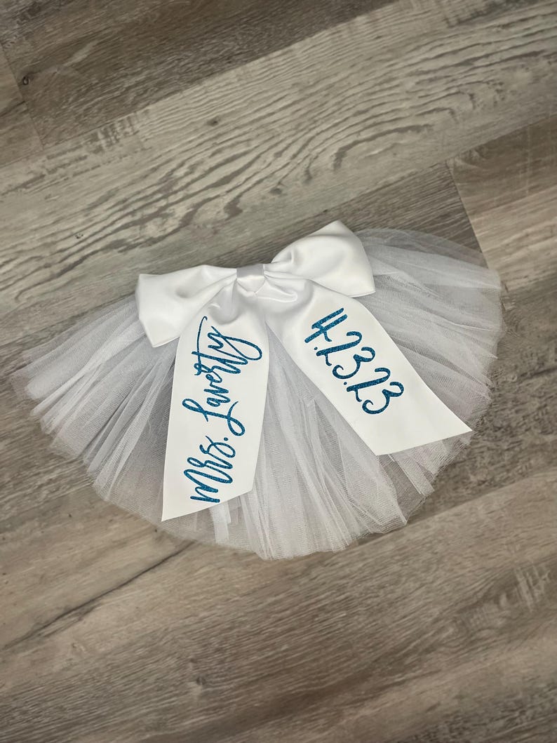 May include: White tulle tutu with a white satin bow and the text "Mrs. Lovely" and "12.3.23" in blue glitter.