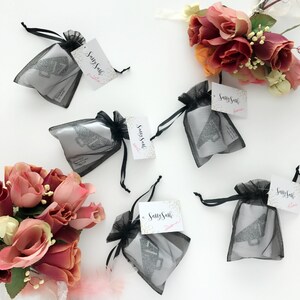 May include: Five black mesh bags with silver glitter megaphones inside. Each bag has a white tag with the text "Sassy Sash" and a pink heart. The bags are on a white surface with a bouquet of pink roses in the lower left corner.
