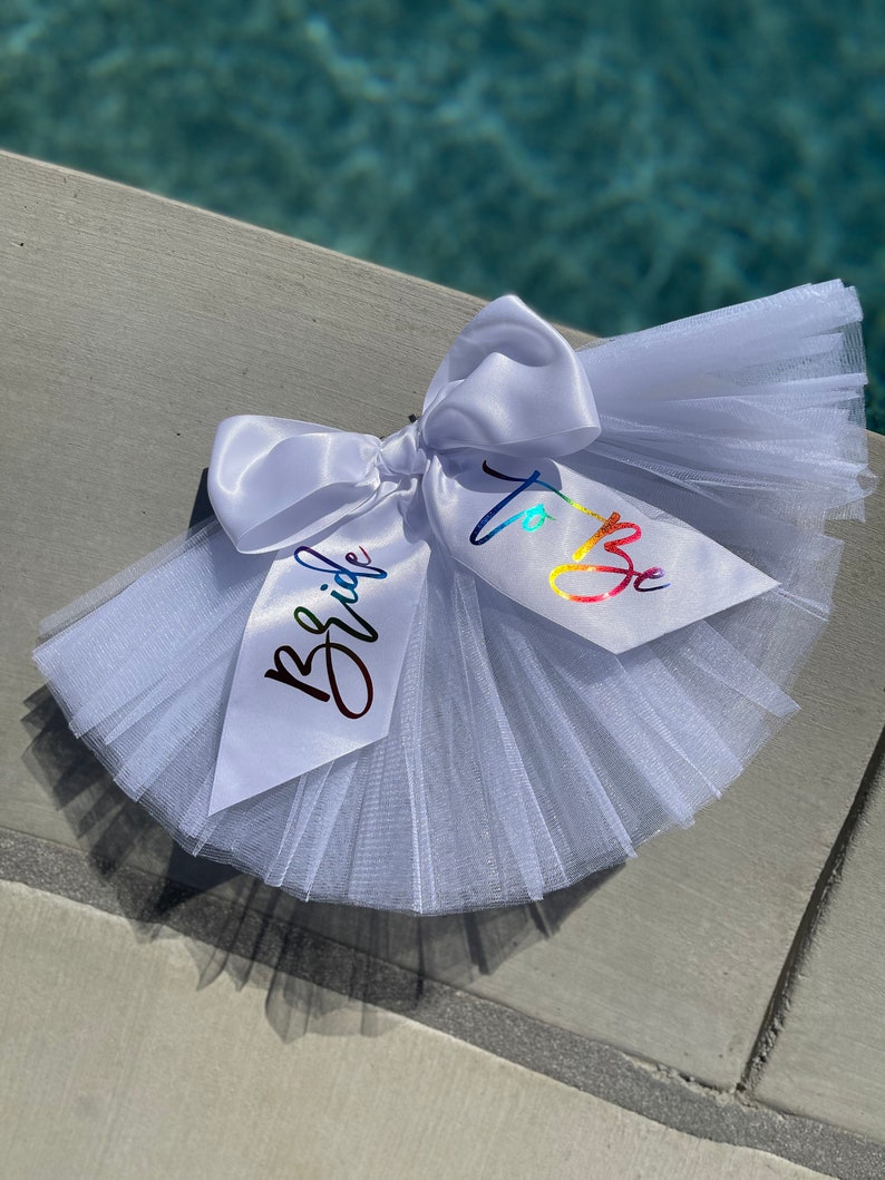 May include: White tulle bridal sash with a white satin bow and rainbow lettering that says "Bride to Be".