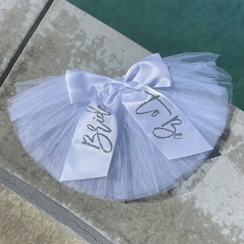 May include: White tulle bridal sash with a white satin bow and silver glitter lettering that reads "Bride" and "To Be".