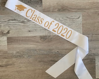 May include: White graduation sash with gold glitter lettering that reads "Class of 2020". A gold glitter graduation cap icon is on the left side of the sash.