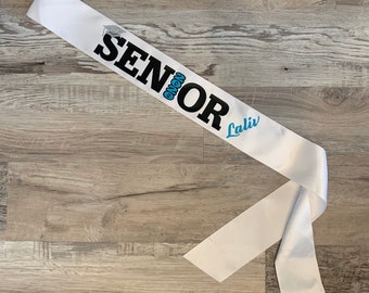 May include: White satin sash with black text that reads "Senior 2023" and blue text that reads "Lali". The sash is tied in a knot and the words "Sassy Sash" are printed on the bottom of the sash.