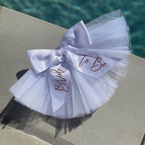 May include: White tulle and satin bow with the words "Bride to Be" written in rose gold glitter.