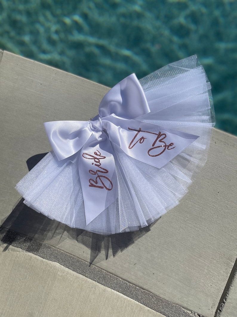 May include: White tulle and satin bow with the words "Bride to Be" written in rose gold glitter.