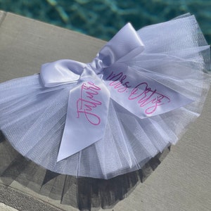 May include: White tulle tutu with a white satin bow and two pink ribbons with the text "Mrs. Ortiz" and "Future" written on them.