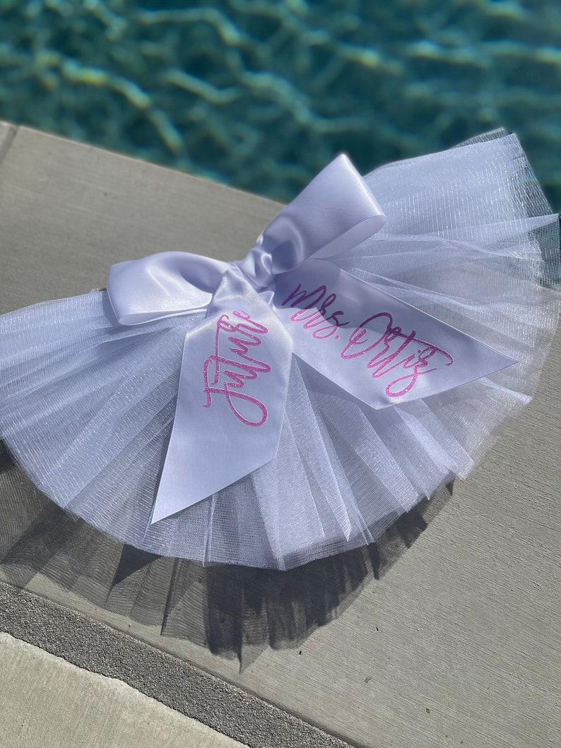 May include: White tulle tutu with a white satin bow and two pink ribbons with the text "Mrs. Ortiz" and "Future" written on them.
