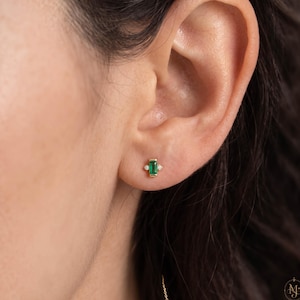May include: A gold stud earring featuring a central rectangular baguette-cut green emerald gemstone flanked by two small round white diamonds on either side. The earring is positioned in the earlobe, showcasing a minimalist metal setting.