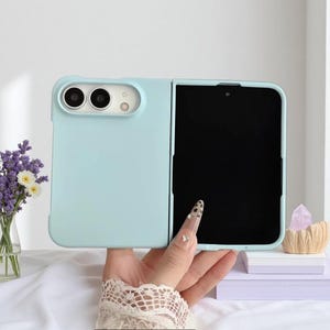 Puede incluir: Una funda para smartphone plegable de color azul claro con un acabado mate suave. La funda presenta un recorte rectangular para un sistema de cámara dual y un flash. El dispositivo se muestra en posición abierta, revelando una pantalla negra grande.