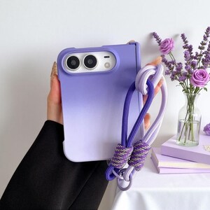 May include: A purple ombre phone case for a foldable smartphone with a dual-lens camera cutout. The case features a detachable, thick woven lanyard strap in matching shades of light and dark purple with metallic-threaded accents at the knots.