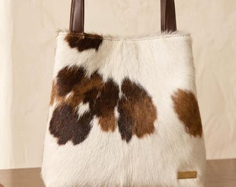 May include: A tote bag featuring a white and brown cowhide pattern with a textured fur finish. The bag includes two dark brown leather handles and a small rectangular gold-toned metal plaque attached to the lower right corner of the front panel.