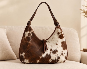 May include: A hobo-style shoulder bag featuring a brown and white cowhide pattern with a soft, textured fur finish. The bag includes a single brown leather strap attached with metal hardware and a central zipper closure with two leather pull tabs.