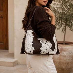 Woman carrying a large cowhide hobo bag on shoulder, styled with brown sweater and white pants for casual outfit