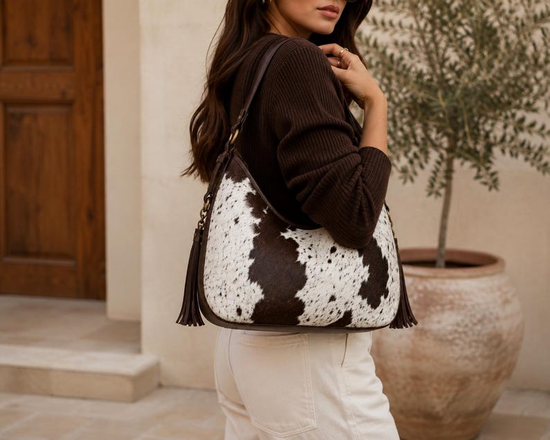 Woman carrying a large cowhide hobo bag on shoulder, styled with brown sweater and white pants for casual outfit