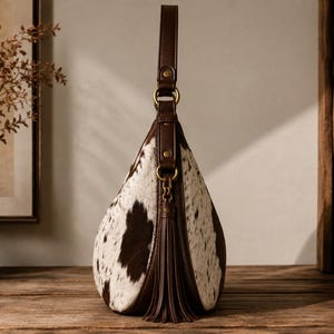 Side view of cowhide leather hobo bag showing rounded silhouette, dark brown leather trim, and fringe tassel detail