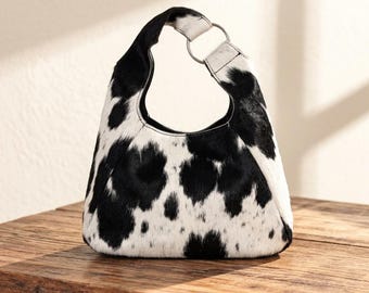 May include: A hobo-style handbag featuring a black and white cow-print pattern with a textured hair-on-hide finish. The bag has a single integrated handle connected by a silver-toned metal ring on the upper right side, creating a curved silhouette.