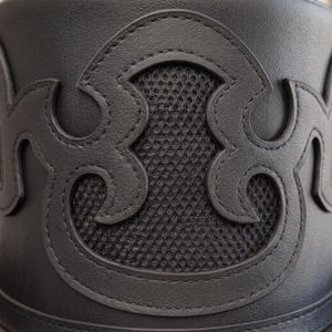 Op de afbeelding: Een zwart cilindervormig object met een glad, leerachtig oppervlak en een opvallende, gestikte, decoratieve overlay in een gebogen, abstracte vorm. Het uitgesneden ontwerp onthult een zwarte mesh-textuur onder het materiaal.