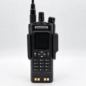 May include: A black Baofeng handheld two-way radio featuring a vertical antenna, a central digital display screen, and a numeric keypad with green lettering. The device is secured within a black plastic belt clip holster and has gold charging contacts.
