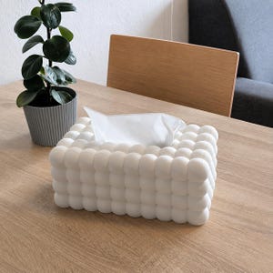 May include: A rectangular white tissue box cover featuring a textured surface composed of numerous small, uniform spheres arranged in a grid pattern. The cover is designed to hold a standard stack of white facial tissues, which protrude from the top.