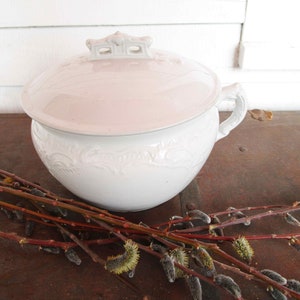 May include: White porcelain chamber pot with a lid and ornate floral design. The pot has a handle on one side and a decorative knob on the lid.