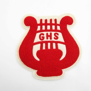 May include: A red and white felt patch in the shape of a lyre. The patch has a textured surface and features the letters "GHS" in white. The lyre shape is outlined in white, with a white background.