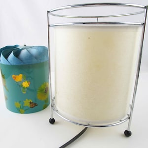 May include: A vintage chrome lamp with a cylindrical off-white shade and a teal container with a floral and butterfly design. The lamp has a curved top and base with black ball feet.