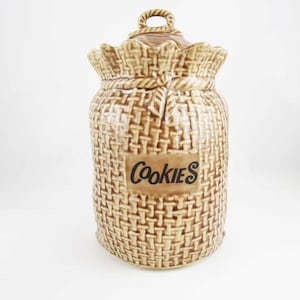 May include: A brown ceramic cookie jar shaped like a burlap sack with a rope handle and the word "Cookies" printed on the front.