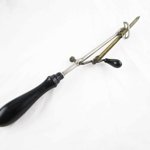 May include: An antique ice shaver with a black wooden handle and metal components. The tool features a blade and a lever, likely used for shaving ice. The metal parts have a silver and brass appearance.