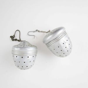 May include: Two silver-colored metal tea infusers shaped like acorns. Each infuser features a perforated body with small circular holes, a rounded cap, and a short metal chain attached to a small hook for securing the device during use.