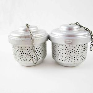 May include: Two silver-colored metal tea infusers with perforated bodies and removable lids. Each infuser features a small metal chain attached to the top, with one chain ending in a hook. The cylindrical containers have a smooth finish.