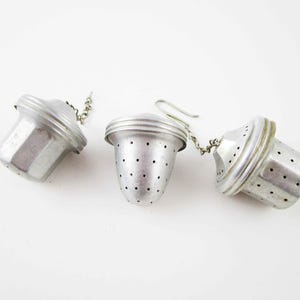 May include: Three silver-toned metal tea infusers with a bell-shaped design. Each infuser features a perforated body for steeping loose leaf tea, a secure lid, and a small attached metal chain with a hook for easy removal from a mug or teapot.