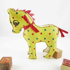 May include: A yellow fabric toy horse with red and blue polka dots and a red ribbon around its neck. The horse has a red fabric tail and is standing on a white surface.