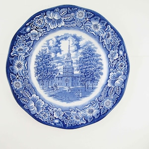 May include: Blue and white decorative plate with a floral border. The center features a detailed illustration of a building with a tall steeple, trees, and figures. The plate has a scalloped edge.