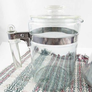 May include: A clear glass coffee pot with a glass lid and a silver metal band around the middle. The handle is clear glass and attached with silver metal. The pot is sitting on a white, red, and green woven mat.