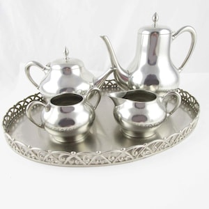 May include: A silver-toned metal tea set consisting of a tall teapot, a shorter teapot, a creamer, and a sugar bowl with lids, all arranged on an oval serving tray with a decorative openwork lattice border and a smooth, reflective surface.