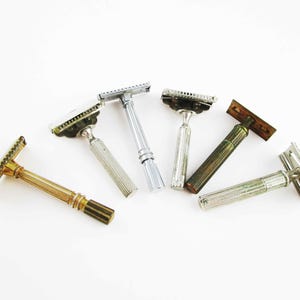 May include: An assortment of vintage safety razors in various metallic finishes, including gold, silver, and bronze. The razors are arranged on a white background, showcasing their classic designs and intricate details. A collection of grooming tools.