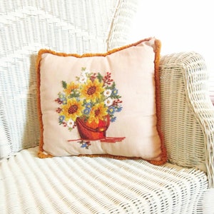 May include: A white wicker chair with a cream-colored pillow featuring an embroidered floral design. The pillow has a brown fringe border.