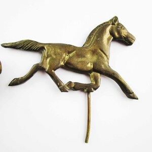 May include: A gold-colored metal horse figurine with a long, thin tail and a raised head. The horse is running and has a long, thin metal rod attached to its underside.