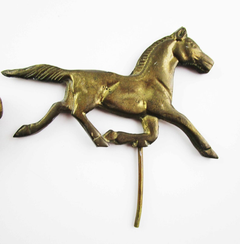 May include: A gold-colored metal horse figurine with a long, thin tail and a raised head. The horse is running and has a long, thin metal rod attached to its underside.