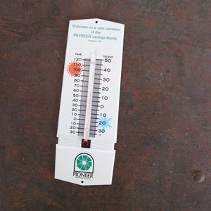 May include: A white thermometer with a red and silver mercury column. The thermometer has a black and white scale with Fahrenheit and Celsius markings. The thermometer is labeled "PIONEER Savings & Trust" and has a sun and snowflake icon.