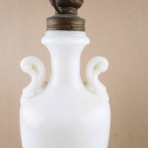 May include: A white, antique-style lamp base with a brass fixture. The lamp has a rounded body with two curved handles. The lamp is made of opaque white glass and has a vintage aesthetic.