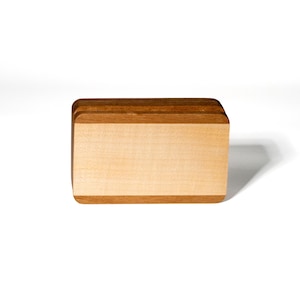 Può includere: Un blocco rettangolare in legno caratterizzato da un pannello frontale di colore chiaro a grana liscia, incorniciato da legno più scuro color bruno-rossastro sui bordi superiore e inferiore. L'oggetto presenta angoli arrotondati e una struttura solida.