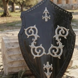Peut inclure: Un bouclier noir en forme d'écu avec une pointe en haut et en bas, orné d'éléments décoratifs en métal argenté. Le bouclier présente une surface lisse et brillante avec un motif géométrique sur les bords et des volutes métalliques symétriques.