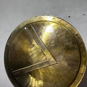 Puede incluir: Un escudo circular de latón con un prominente símbolo lambda en forma de V en relieve en el centro. La superficie metálica tiene un acabado pulido y reflectante con texturas martilladas visibles y remaches en el borde exterior.