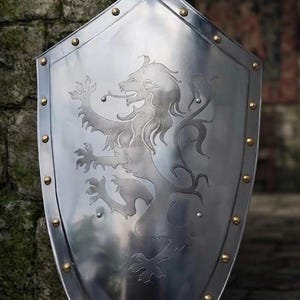 May include: A polished silver-toned metal heater shield featuring a central engraved rampant lion design. The shield has a pointed base and is bordered by a series of evenly spaced, small, round brass-colored rivets along the entire outer edge.