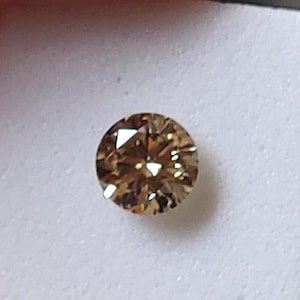 May include: A round, brilliant-cut gemstone with a warm, light brown hue. The stone features multiple facets that reflect light, creating a sparkling appearance. It is securely mounted on a small, square, white foam display card for presentation.