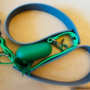 May include: A gray and green dog leash with a poop bag dispenser attached. The leash is made of a durable material and has a metal clip.